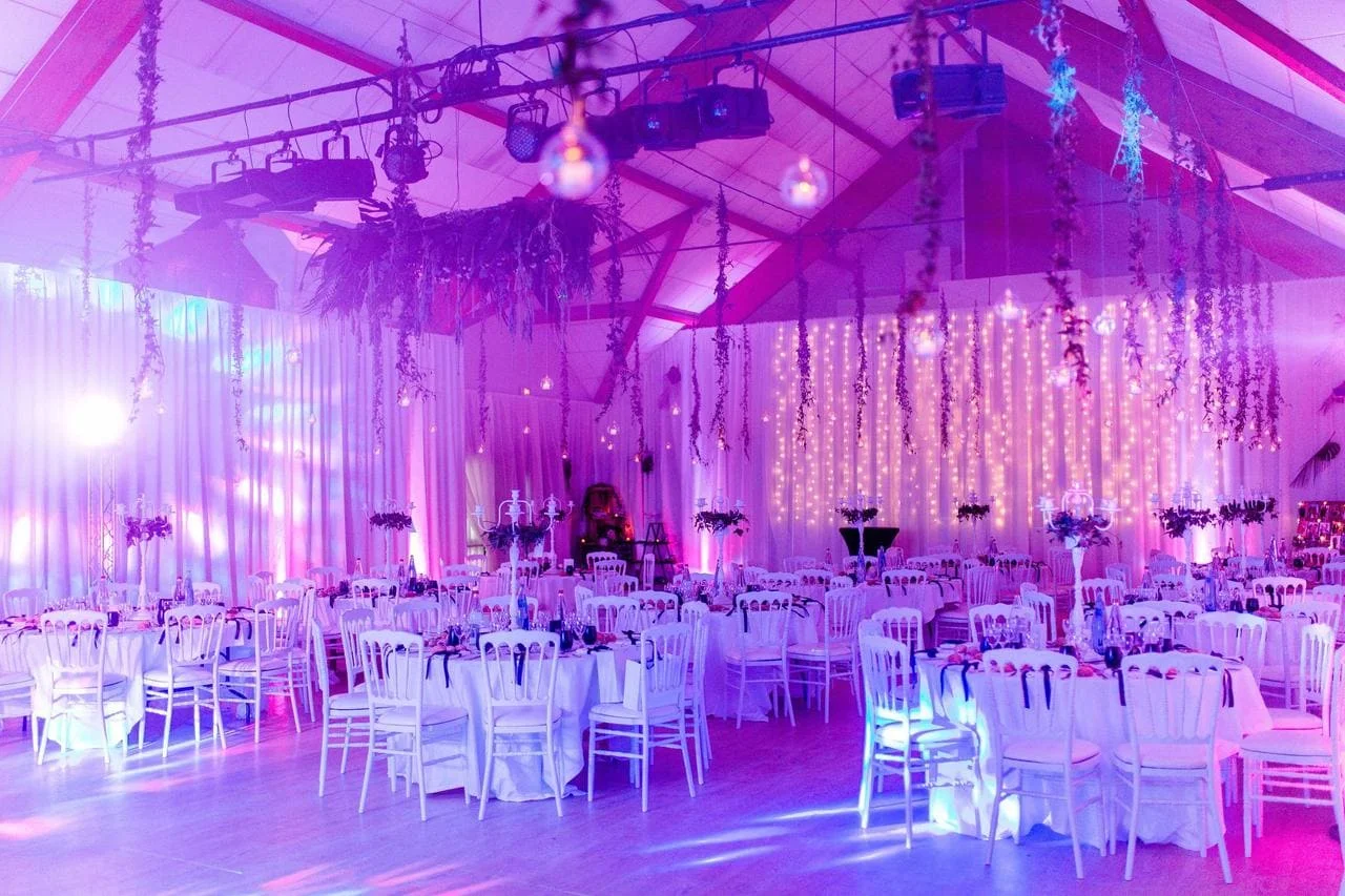 Une salle de fêtes éclairée par une lumière violacée avec des tables blanches et nappes blanches